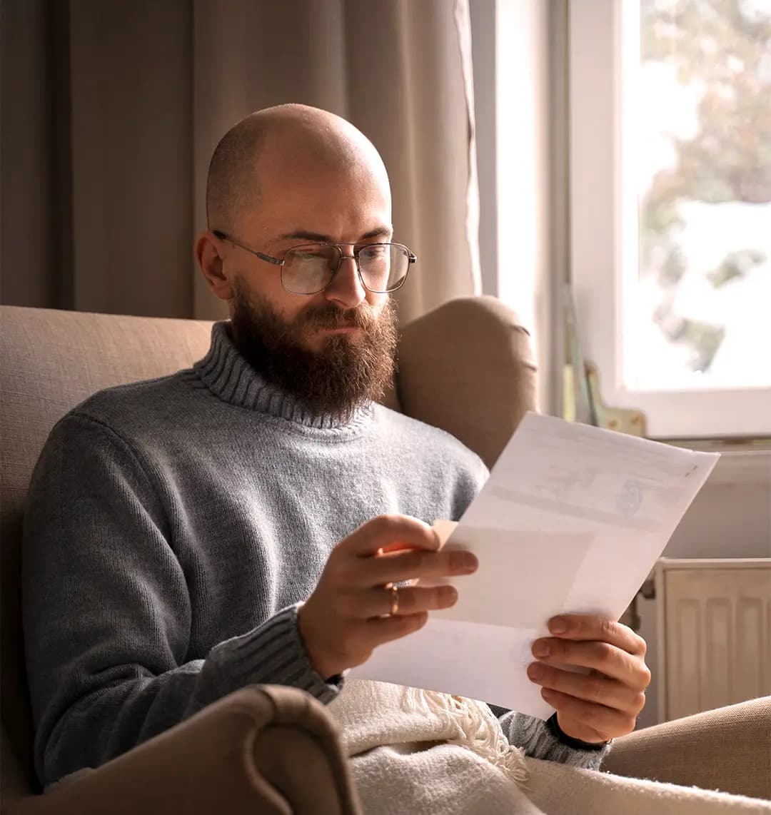 Ein bärtiger Mann mit Brille liest ein Dokument, während er in einem Sessel sitzt, eingewickelt in eine Decke in der Nähe eines Fensters.