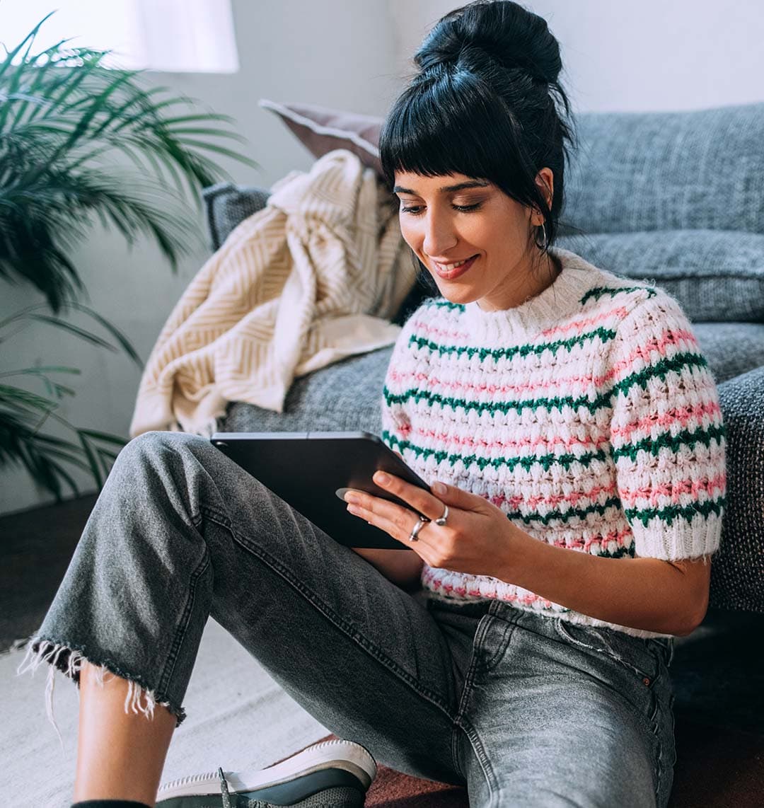 Eine Frau sitzt im Fokus des Bildes und schaut auf ein Tablet, dass sie in den Händen hält. Sie befindet sich in einem Raum und lehnt sich an ein Sofa.
