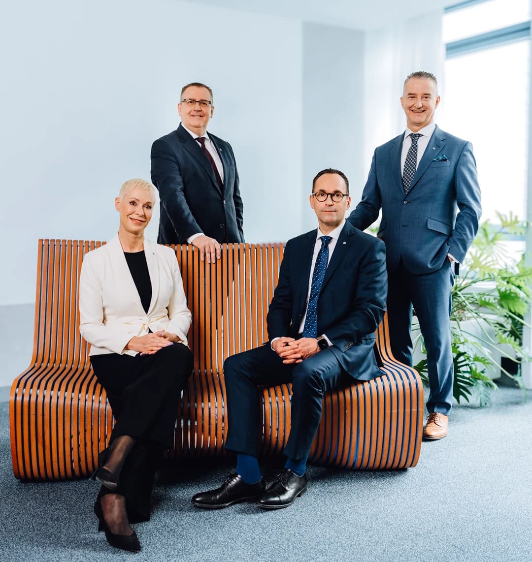 Vier Vorstandsmitglieder in Businesskleidung posieren in einem modernen Büro, zwei sitzen auf einer Holzbank, zwei stehen dahinter.