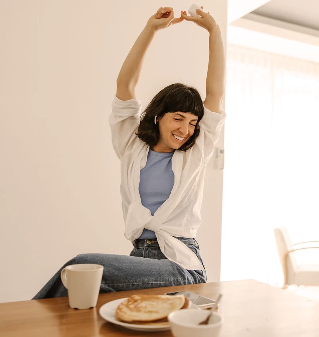 Eine dunkelhaarige Frau sitzt an einem Tisch. Sie lächelt und streckt die Hände in die Höhe. Vor ihr auf dem Tisch sind eine Kaffeetasse, Pancakes und eine Zuckerschale. Die Stimmung ist warm und entspannt.
