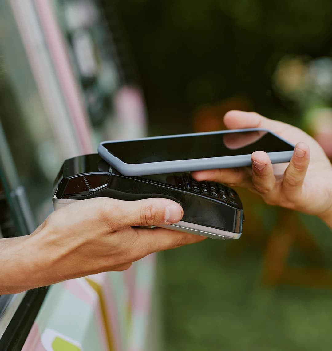 Person, die im Freien mit einem Smartphone über ein von einer anderen Person gehaltenes Kartenlesegerät eine kontaktlose Zahlung vornimmt.