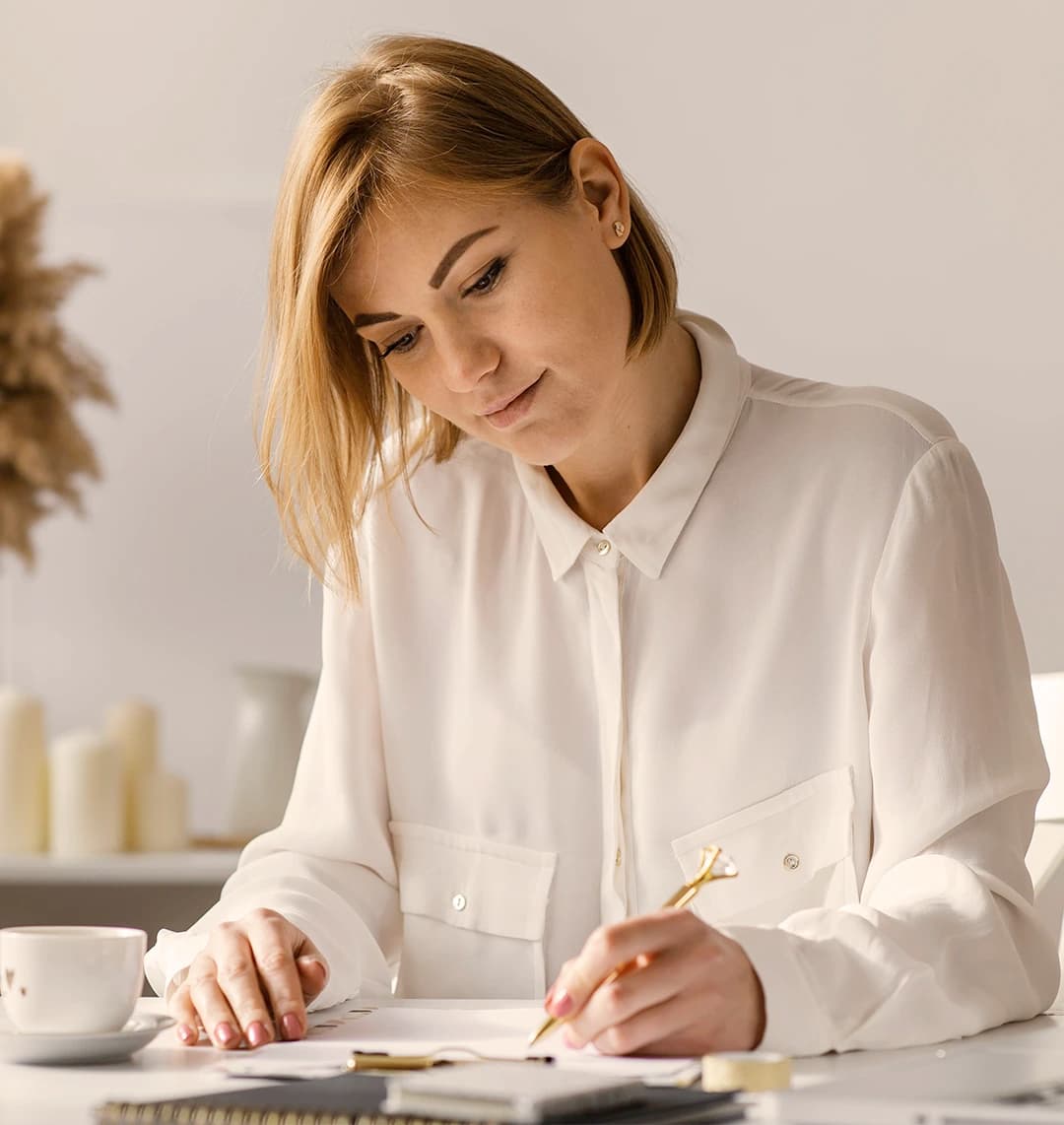 Eine junge Frau mit Pagenschnitt sitzt am Tisch. In der linken Hand hält sie einen goldenen Kugelschreiber und macht Notizen. Rechts vor ihr steht eine weiße Kaffeetasse.