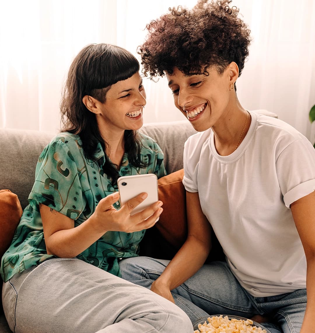 Zwei junge Frauen schauen gemeinsam lachend auf dem Sofa in ein Smartphone.