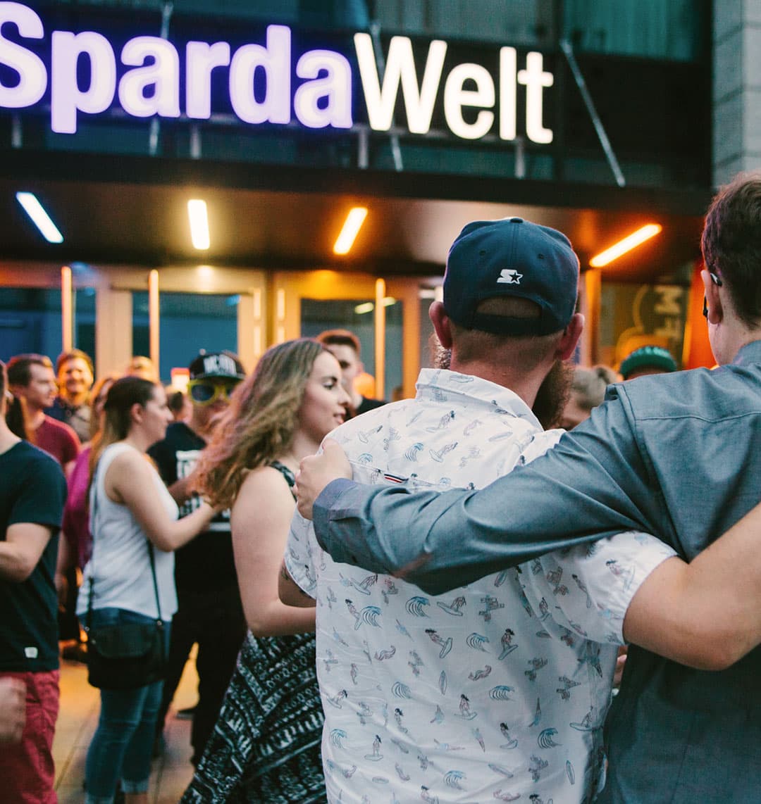 Auf dem Bild ist eine Menschenmenge vor dem Eingang eines Gebäudes, dem SpardaWelt Eventcenter zu sehen. Im Vordergrund sind zwei Personen zu sehen, die ihre Arme umeinander legen.