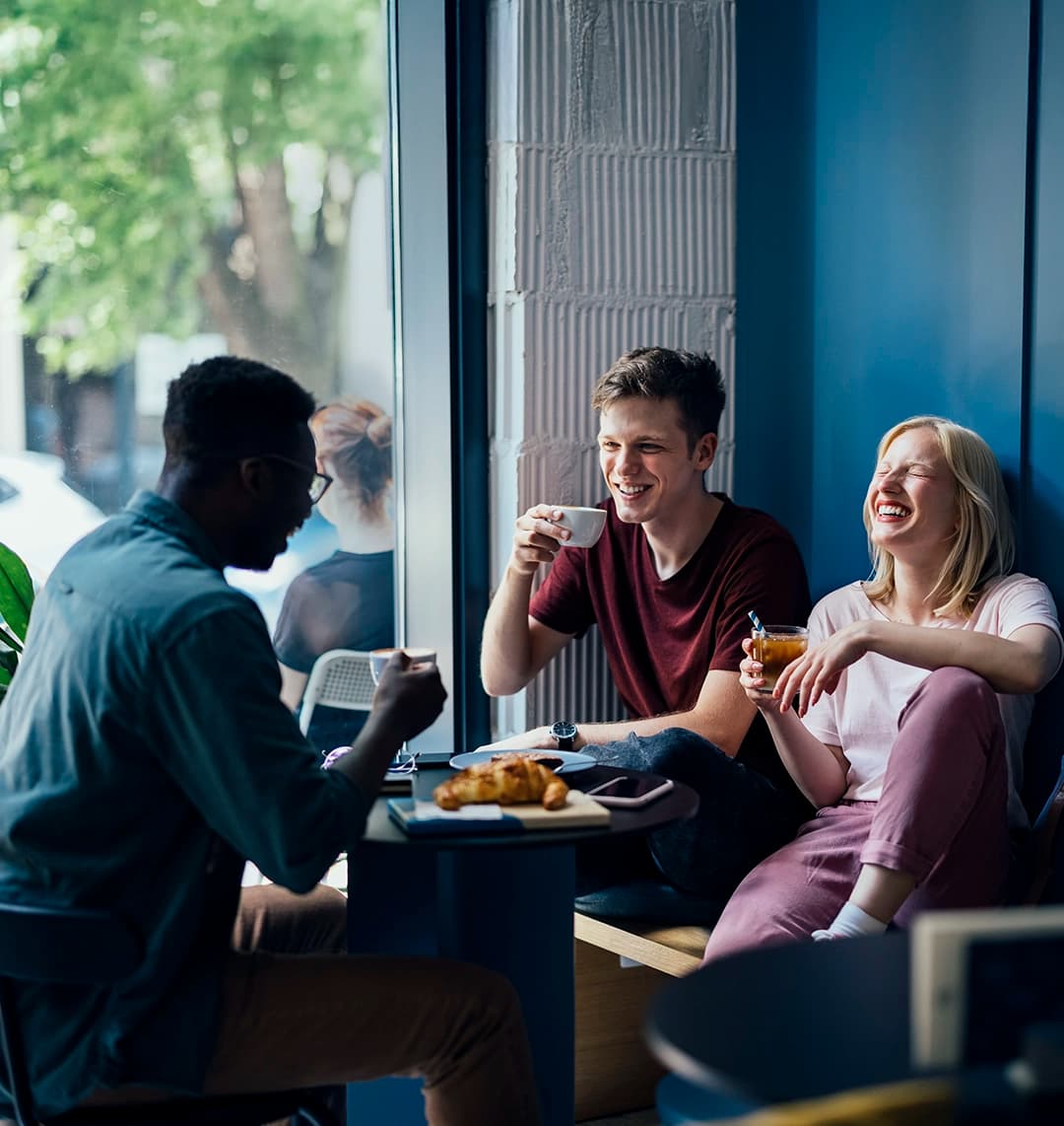Drei junge Erwachsene sitzen lachend und entspannt in einem modernen Café an einem Tisch am Fenster und genießen gemeinsam Kaffee und Gebäck.