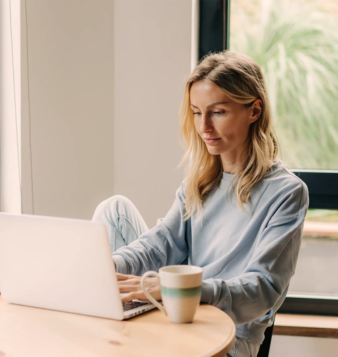 Eine Frau in einem leichten Pullover sitzt an einem Tisch und tippt auf einem Laptop. Daneben steht eine Tasse, in einem hellen Raum mit einem großen Fenster.