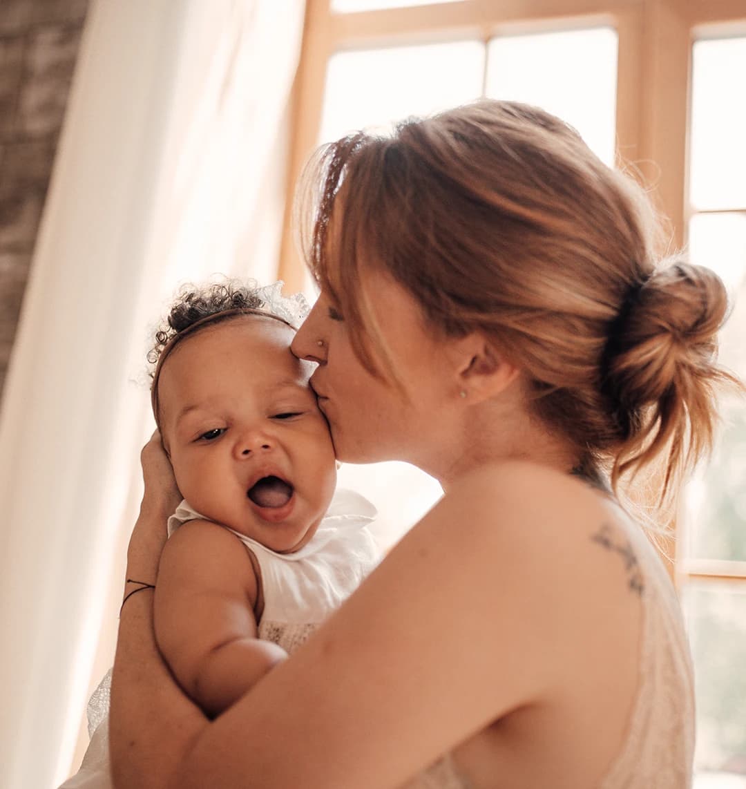 Eine Frau hält ihr Baby im Arm und küsst es sanft auf den Kopf. Das Baby gähnt müde in die Kamera.