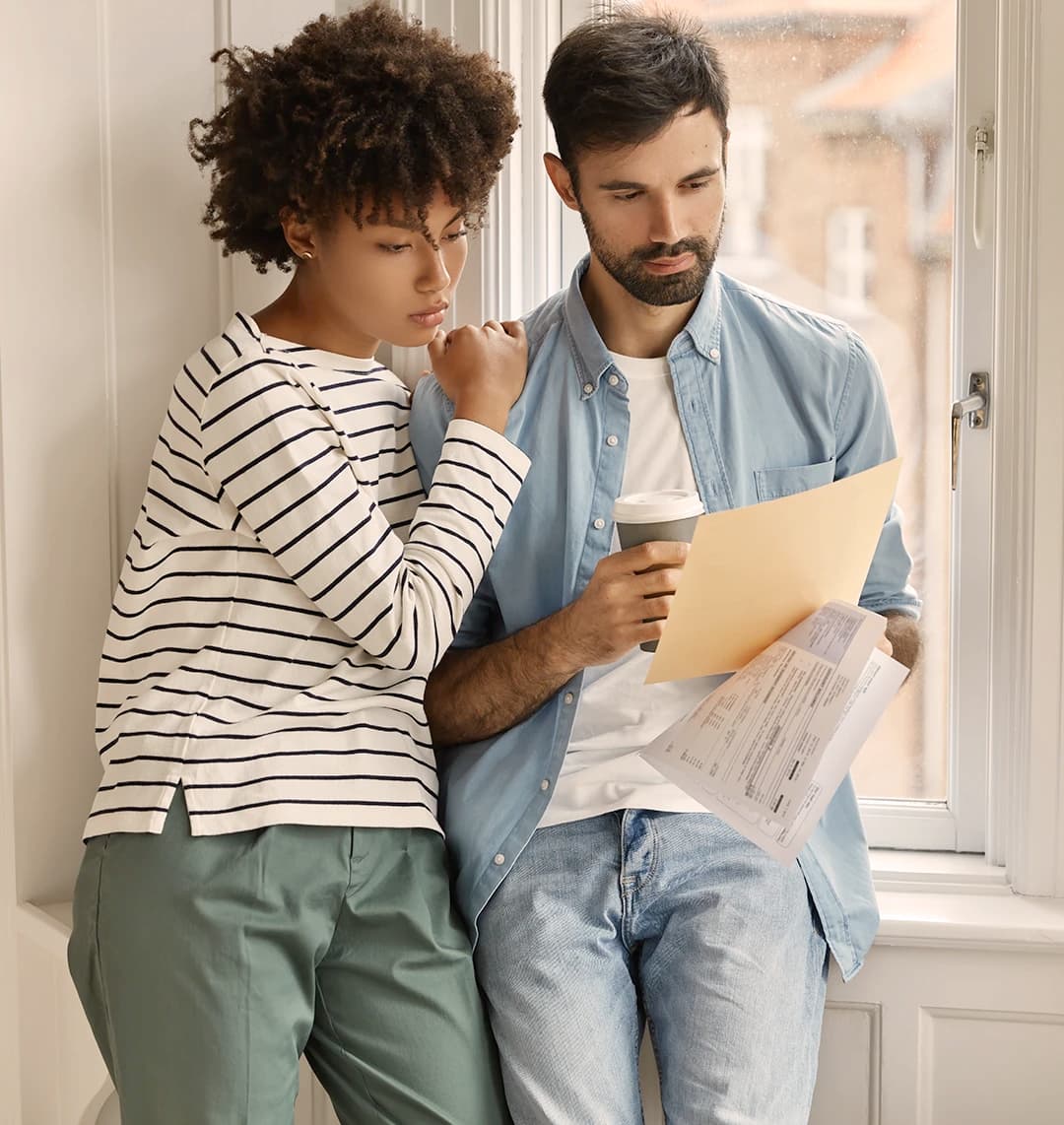 Ein junges Paar lehnt an einer Fensterbank. Sie schauen gemeinsam auf Unterlagen, die er in seiner linken Hand hält. Beide schauen ernst und interessiert. Sei lehnt sich an seine rechte Seite.