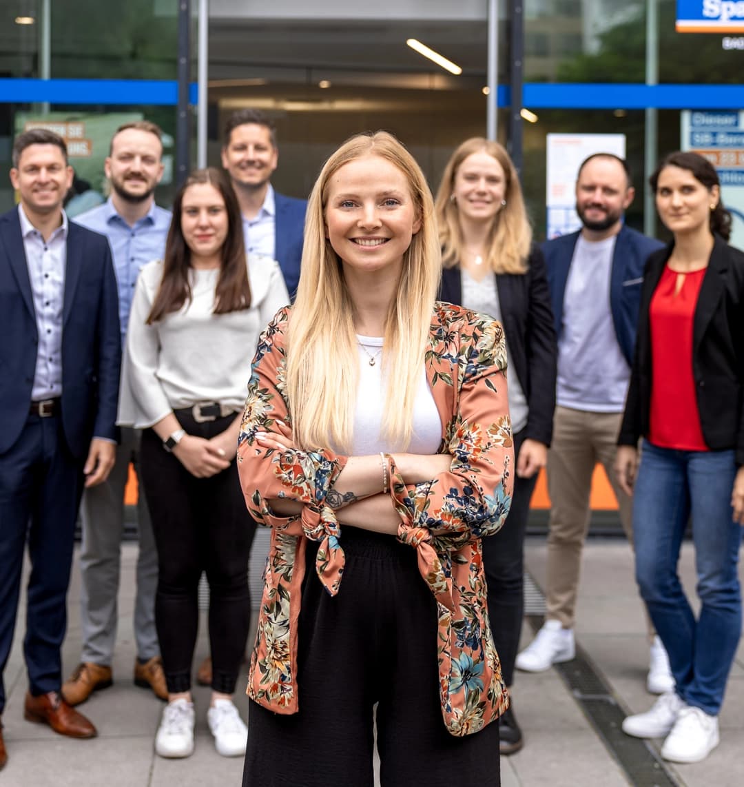 Eine junge Frau steht selbstbewusst im Vordergrund, während acht Kolleginnen und Kollegen lächelnd im Hintergrund vor einem modernen Gebäude posieren