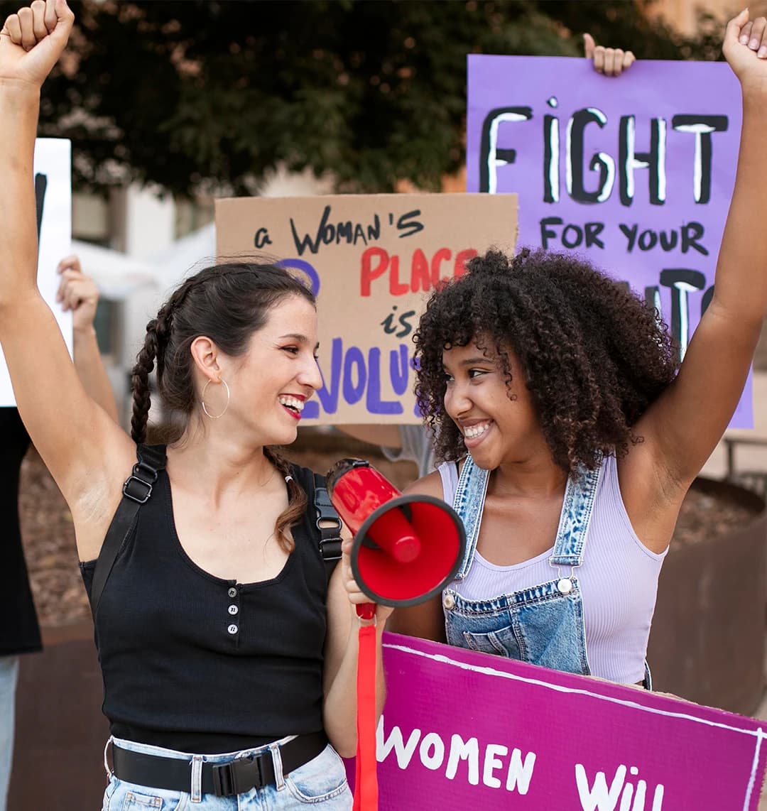 Eine Gruppe demonstriert friedlich für die Rechte von Frauen. Zwei junge Frauen halten ein Mikro in der Hand. Sie lächeln sich glücklich an und strecken ihre Arme in die Luft.
