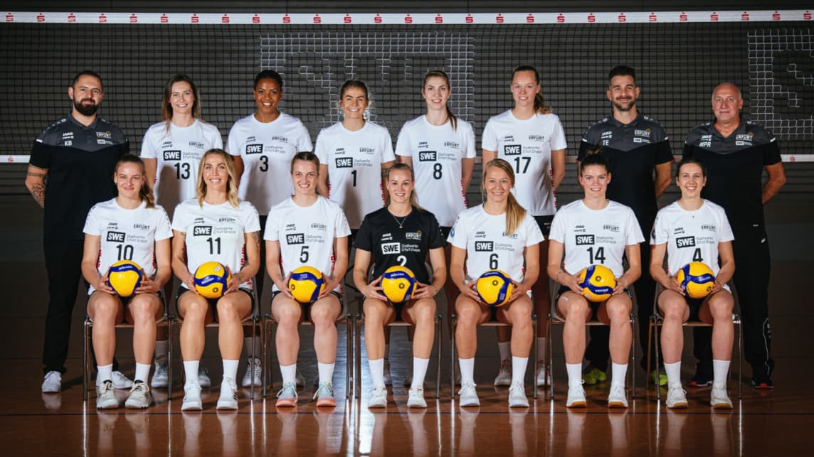 Eine Frauenvolleyballmannschaft posiert in Uniform mit Trainern vor einem Netz.
