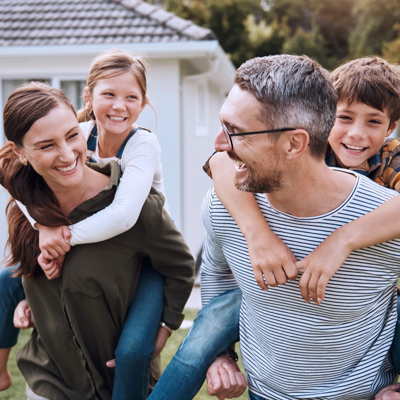 Ein Mann und eine Frau stehen vor einem Haus und tragen jeweils ein Kind huckepack. Alle lachen, der Mann und die Frau schauen sich dabei an.
