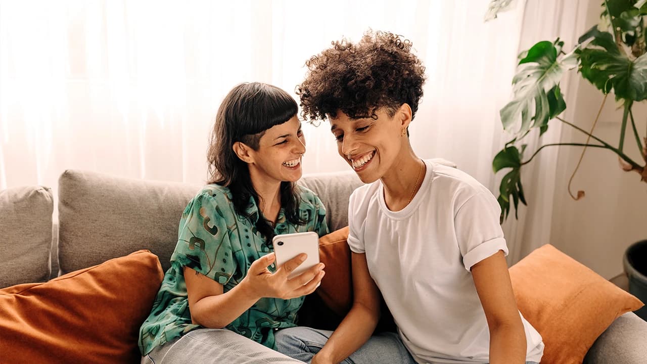 Zwei junge Frauen schauen gemeinsam lachend auf dem Sofa in ein Smartphone.