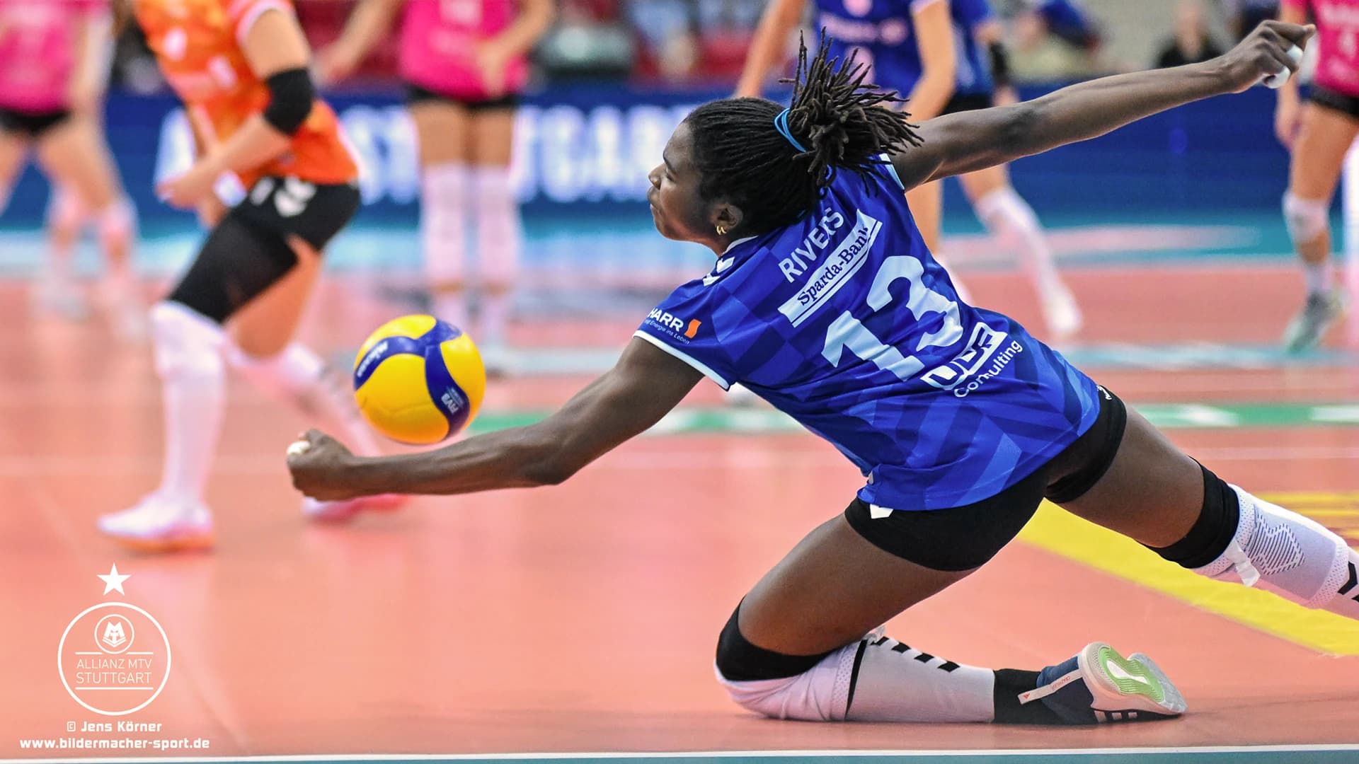 Eine Volleyballspielerin im blauen Trikot mit der Nummer 13 stürzt sich auf einen Hallenplatz, um den Ball zu schlagen.