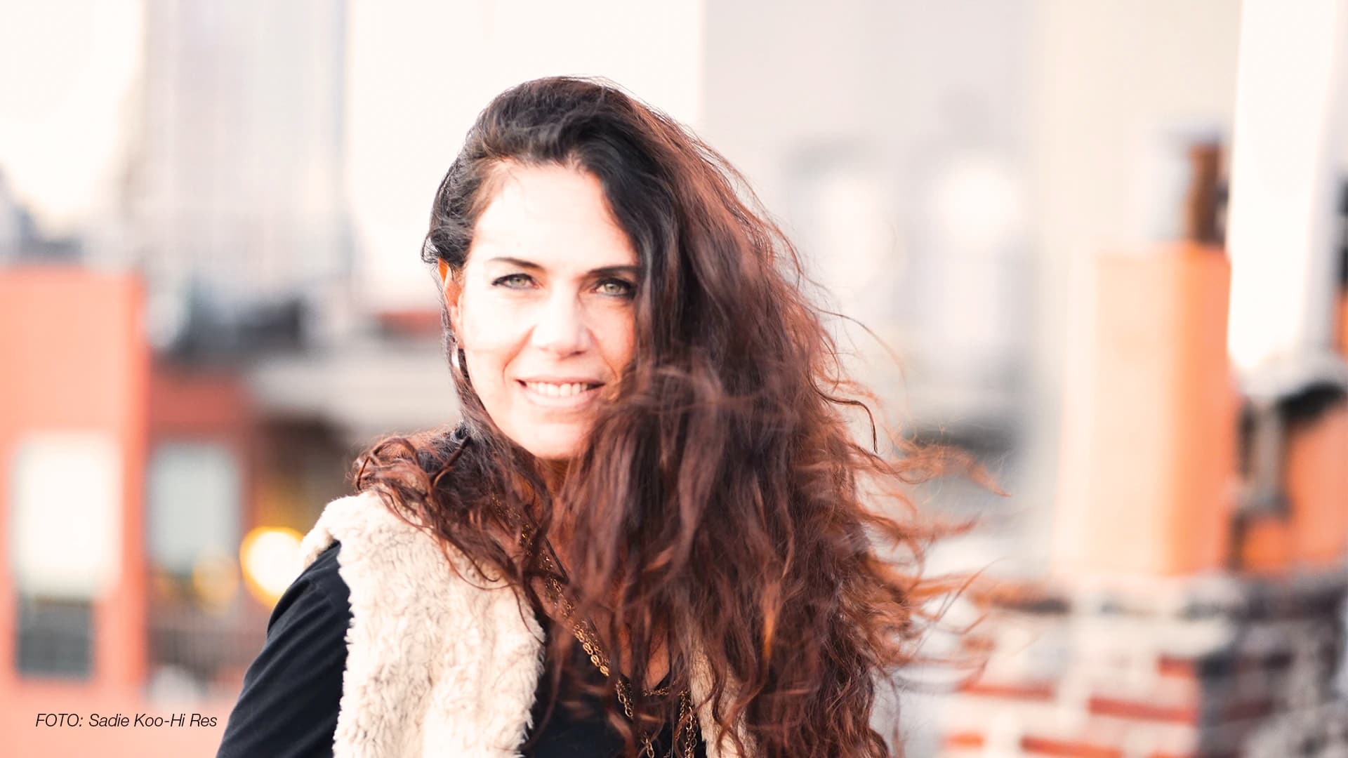 Sari Schorr steht im Fokus des Bildes, Wind weht durch ihre langen braunen Locken, sie lächelt leicht in die Kamera.