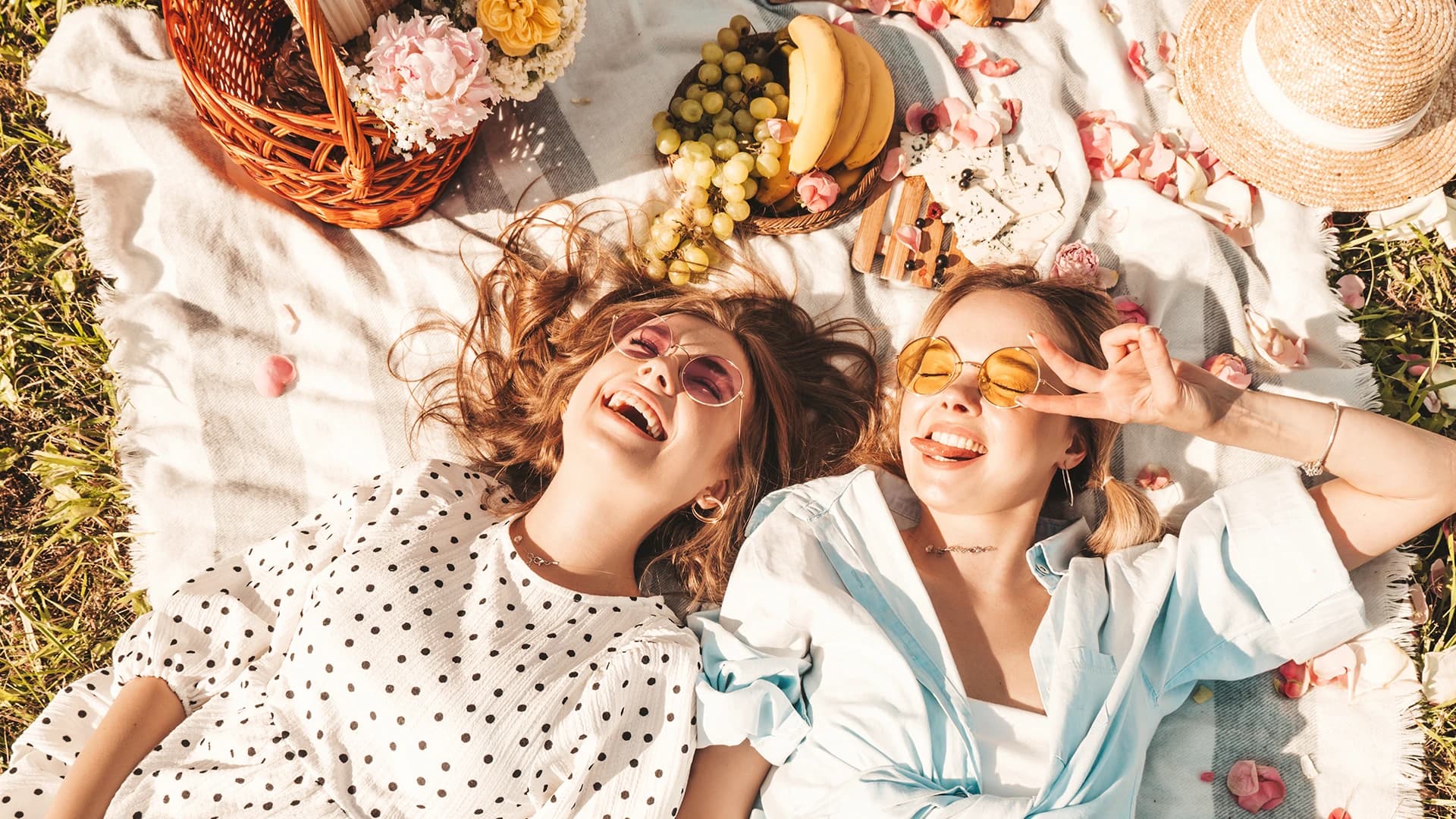 Zwei Frauen liegen lachend auf einer Picknickdecke, umgeben von Früchten, Blumen und einem Weidenkorb, daneben stehen eine Sonnenbrille und ein Sonnenhut.