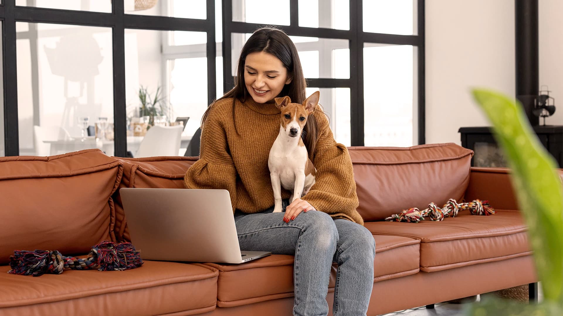 Eine junge Frau sitzt auf ihrem Sofa, mit einem Hund auf dem Schoss. Sie blickt lächelnd in den Laptop vor ihr.