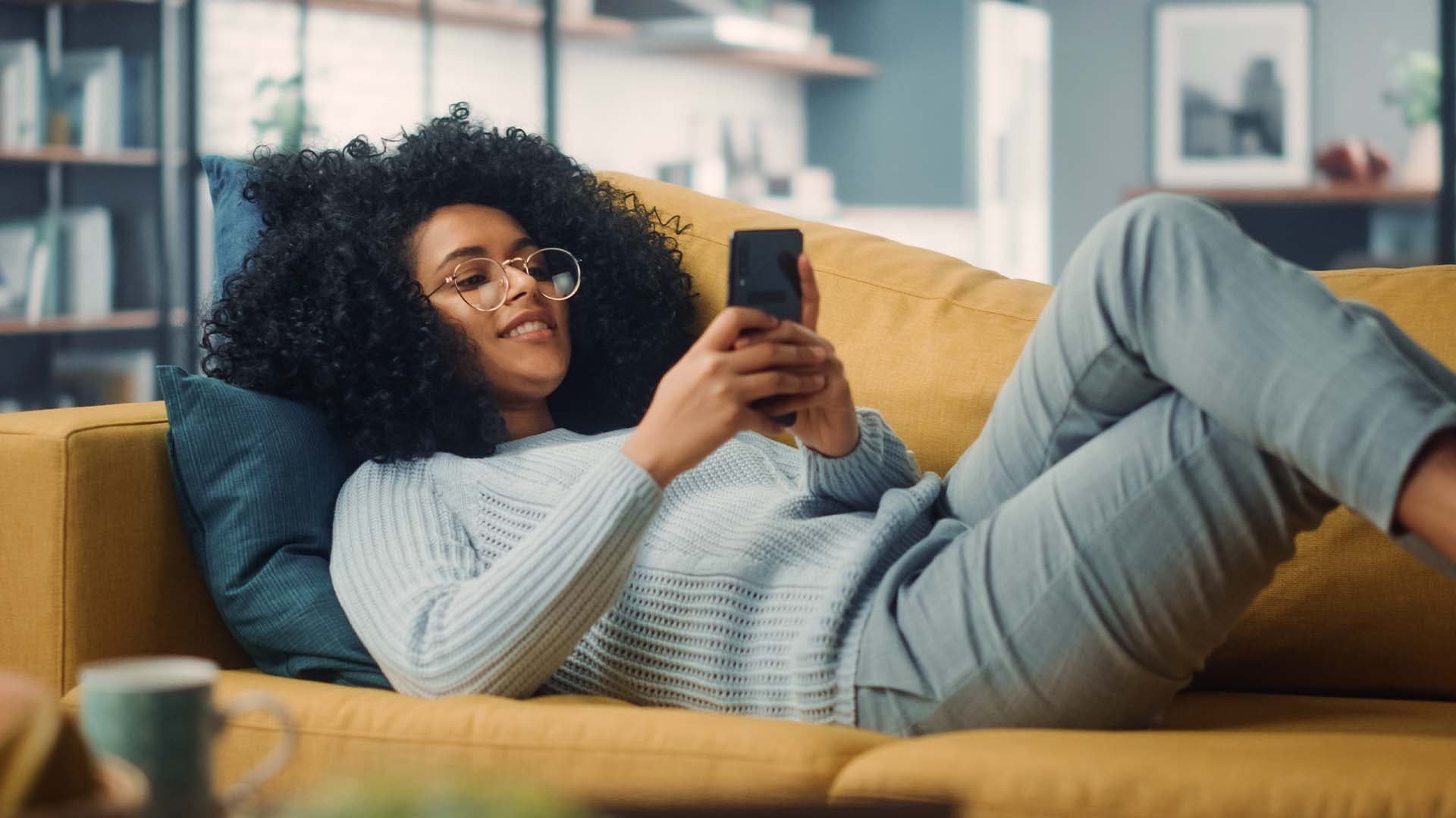 Eine junge Frau liegt auf der Couch und liest an ihrem Handy den Banking Duden der Sparda BW.
