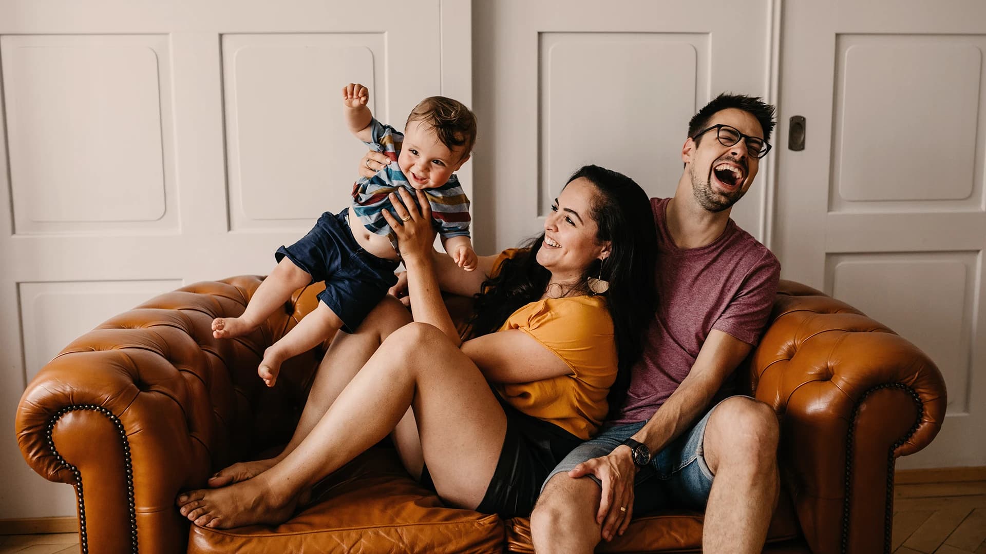 Eine fröhliche Familie sitzt auf einem braunen Ledersofa; die Mutter hält ein lächelndes Baby und der Vater lacht neben ihnen.