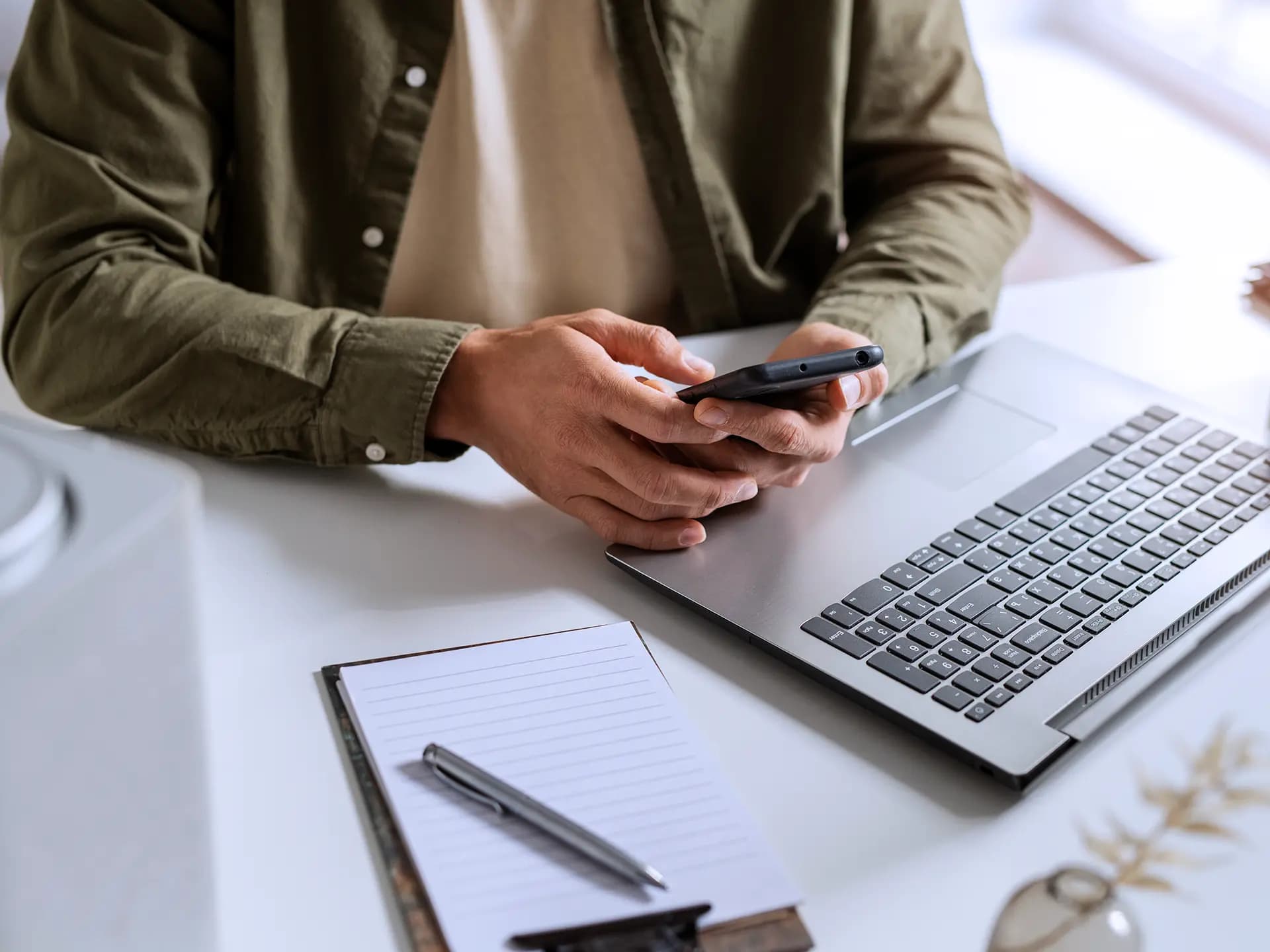 Eine Person sitzt an einem Laptop mit einem Handy in der Hand. Daneben auf dem Tisch liegen Stift und Papier.