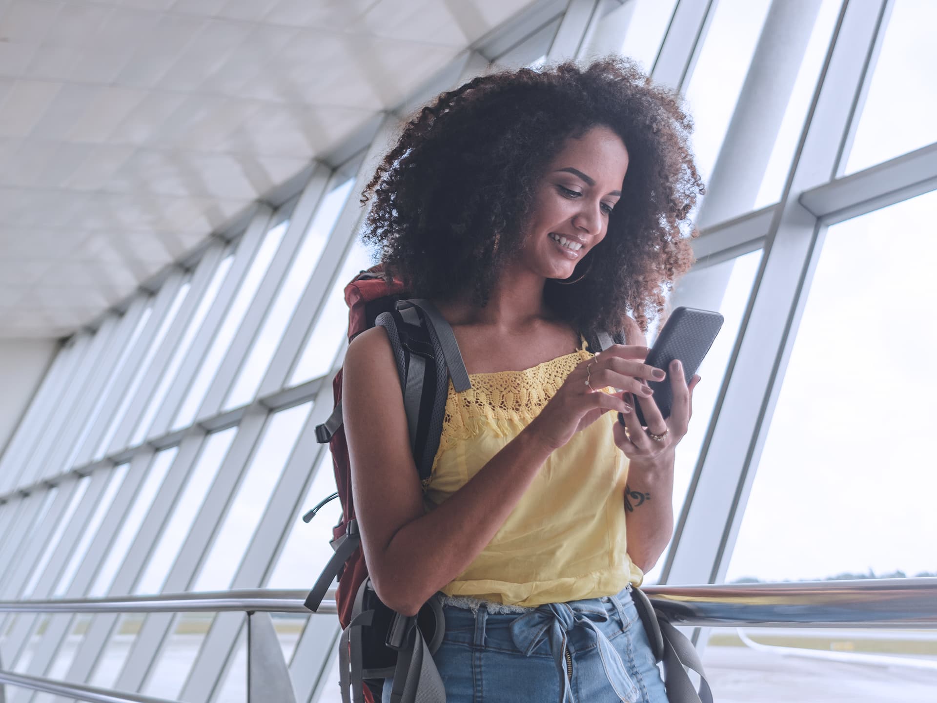 Eine Frau steht mit einem Smartphone in der Hand vor einer Glasfront in einem Flughafen. Sie trägt einen Rucksack auf dem Rücken und lächelt, während sie in das Smartphone schaut.