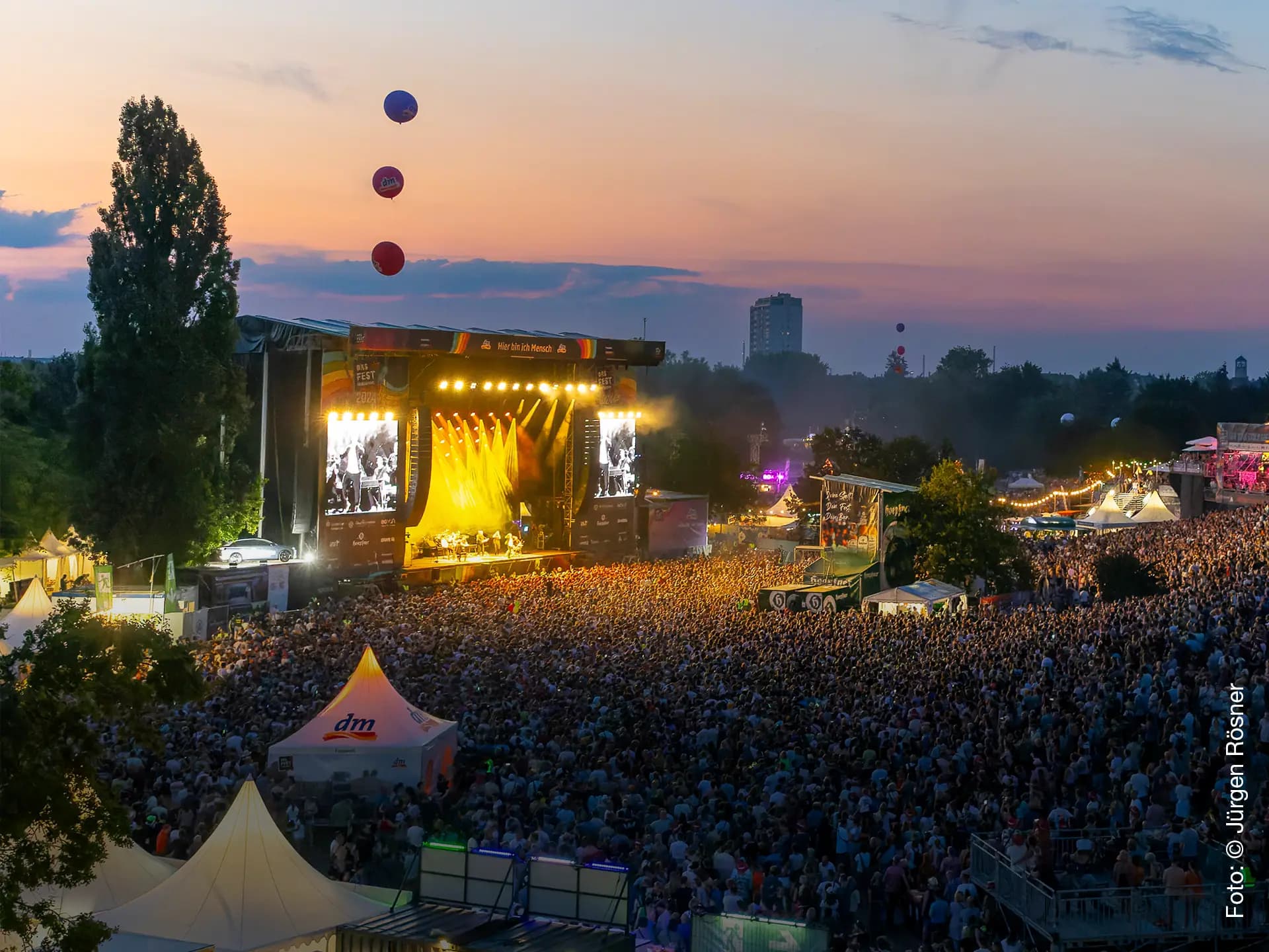 Luftaufnahme des DAS FEST Festivals im Freien bei Sonnenuntergang, mit einer Menschenmenge, der beleuchteten Hauptbühne am Mount Klotz und Luftballons am Himmel.