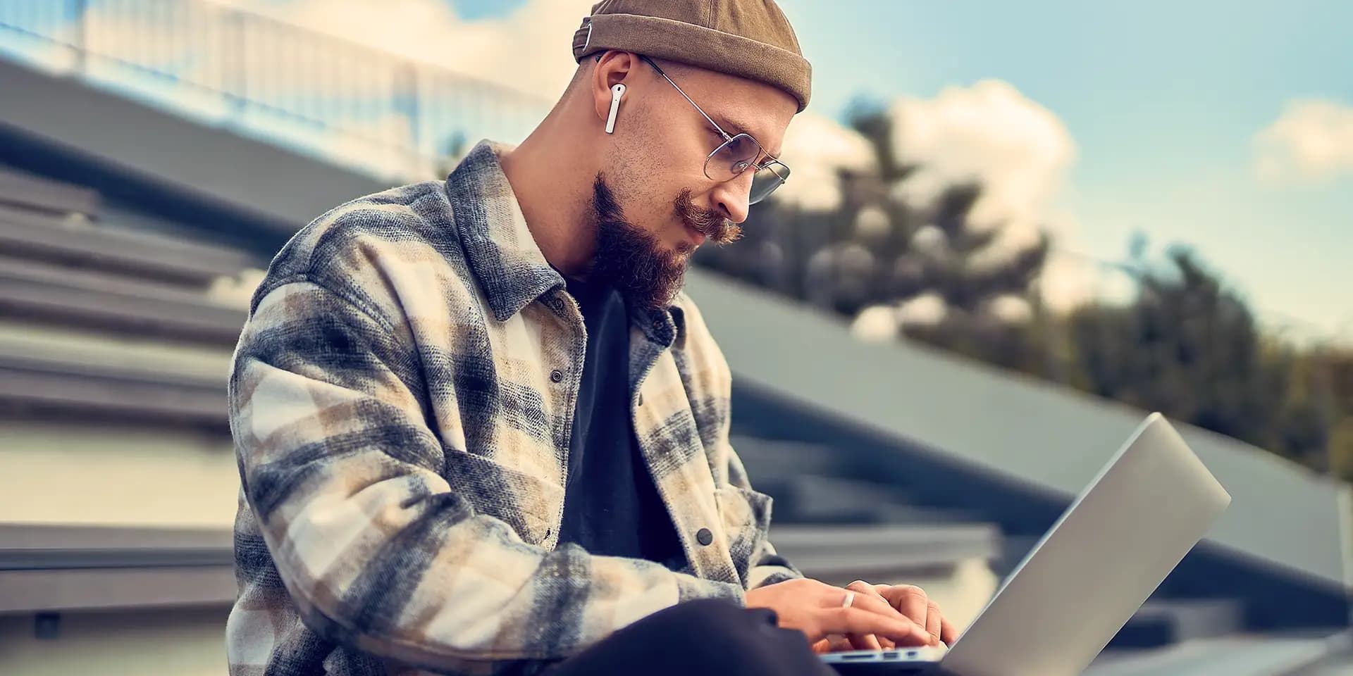 Mann mit Brille und Mütze, der im Freien einen Laptop benutzt, mit karierter Jacke und kabellosen Kopfhörern, mit unscharfen Schritten und Himmel im Hintergrund.