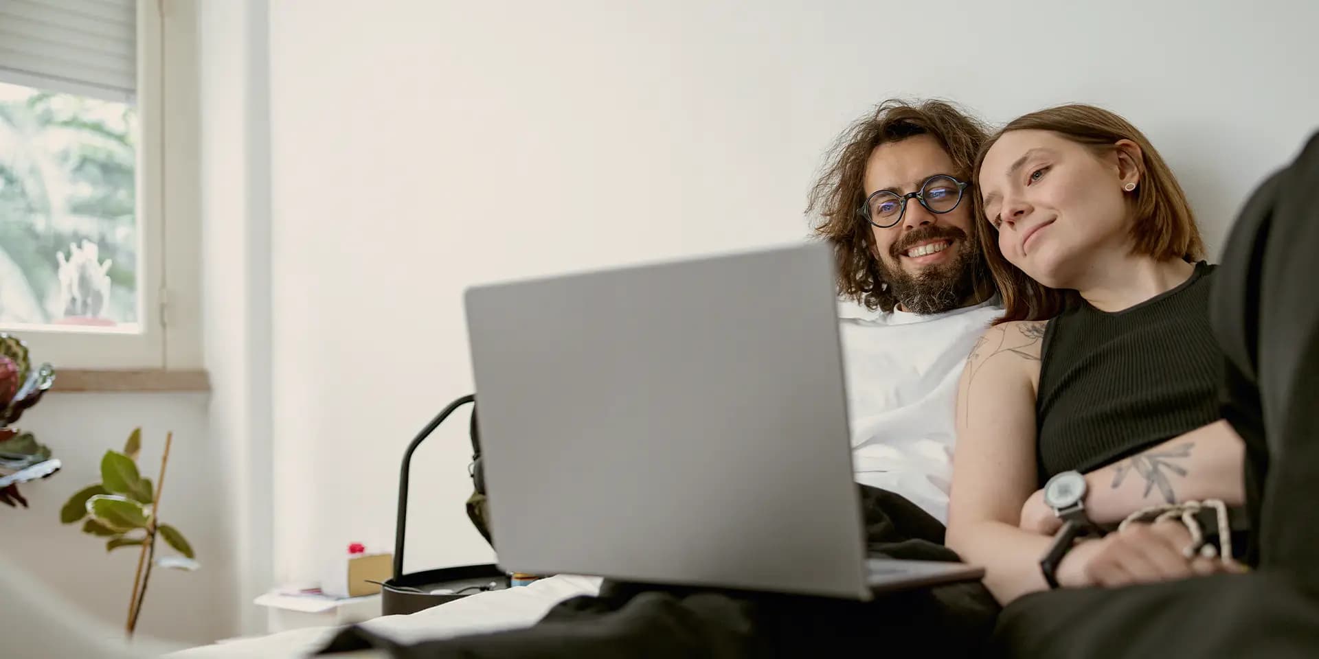 Ein Paar sitzt auf einem Sofa und schaut fröhlich in einen Laptop, der bei dem Mann auf dem Schoß sitzt.