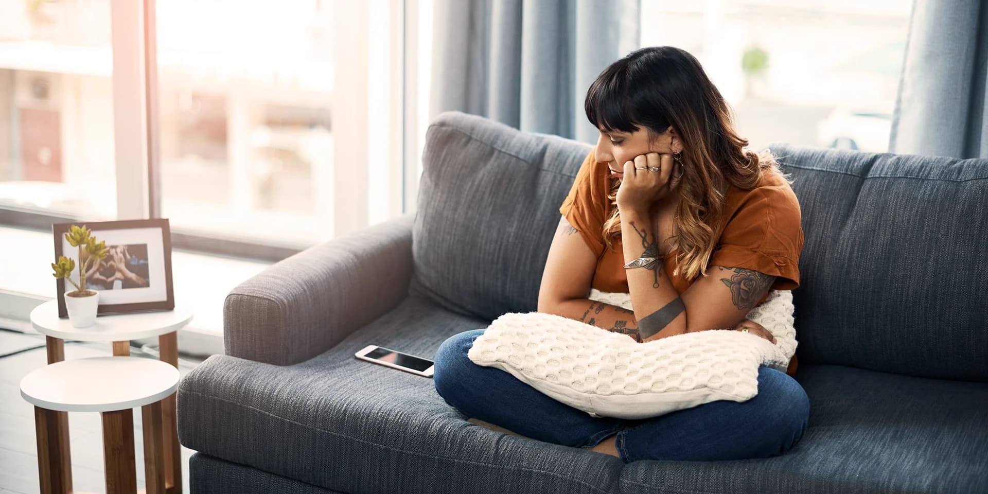 Eine Frau sitzt in ihrem gemütlichen Wohnzimmer auf dem Sofa und blickt traurig auf das Handy neben ihr. Sie stützt ihren Kopf mit ihrer Hand, auf ihrem Schoß liegt ein Kissen.