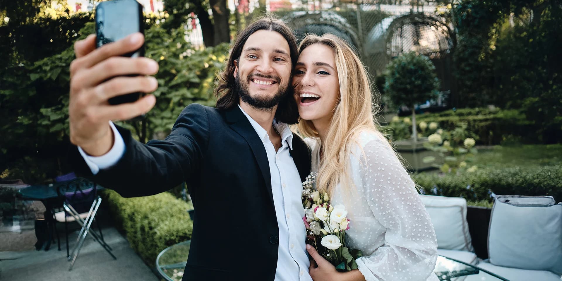 Ein junges Hochzeitspaar steht vor einem grünen Garten. Beide lächeln glücklich und machen ein Selfie mit dem Handy.