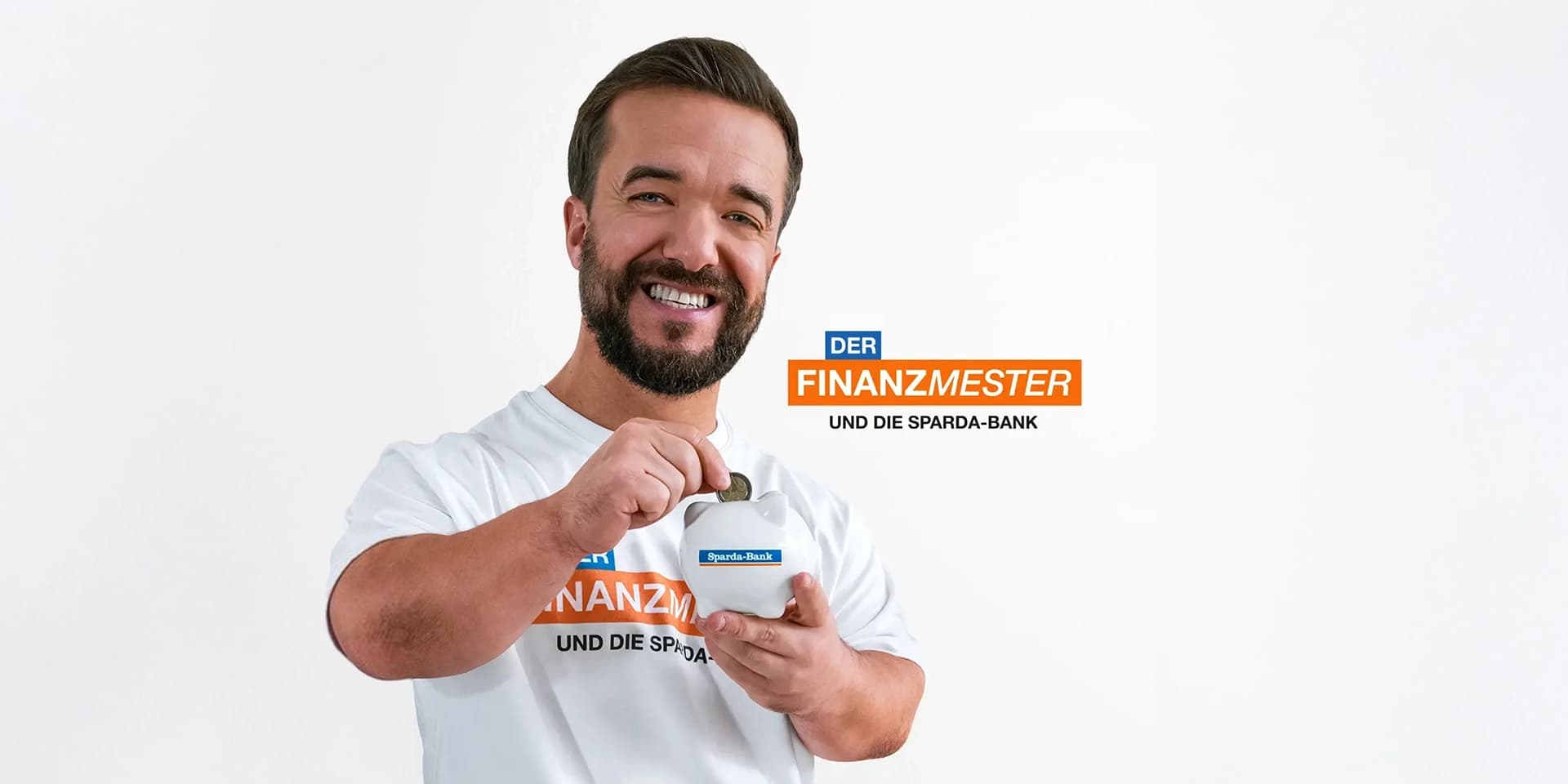 Ein lächelnder Mathias Mester steckt eine Münze in ein Sparschwein mit Sparda-Bank-Logo und trägt ein T-Shirt mit „DER FINANZMESTER UND DIE SPARDA-BANK“