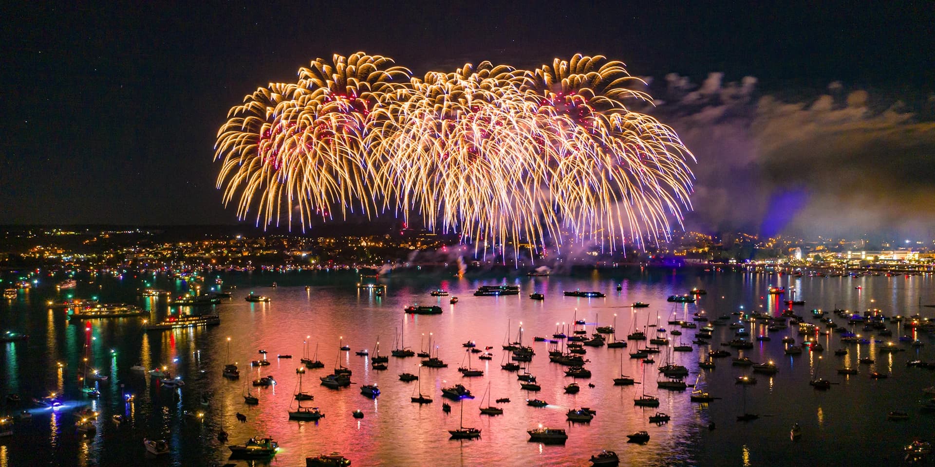 Auf dem Bodensee schwimmen viele kleine Boote, im Hintergrund ist am dunklen Himmel ein buntes Feuerwerk.