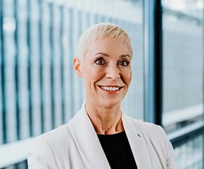 Susanne Drescher mit kurzen blonden Haaren und einem weißen Blazer lächelt vor einem großen Fenster.