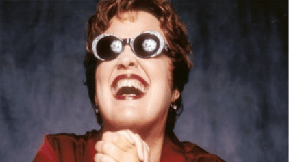 Fröhlich lachende Person mit Sonnenbrille mit reflektierendem Muster, dunklem Lippenstift und rotem Oberteil vor dunklem Hintergrund.