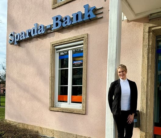Eine Frau im schwarzen Anzug steht lächelnd vor einem Gebäude mit einem „Sparda-Bank“-Schild.