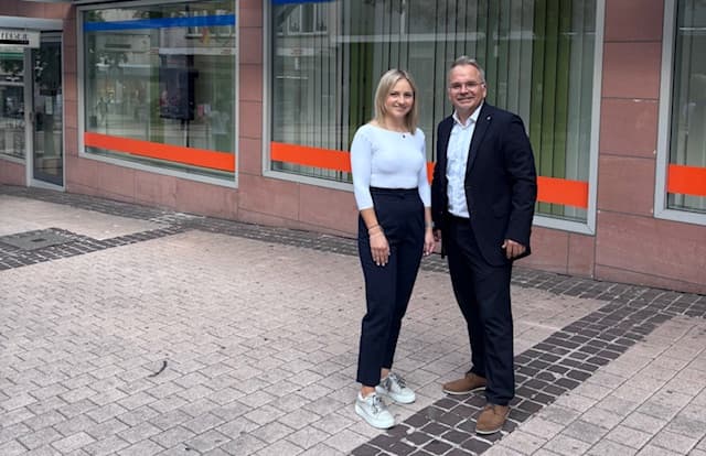 Eine Frau und ein Mann stehen auf einem Bürgersteig vor einem Gebäude mit großen Fenstern und orangefarbenen Akzenten.