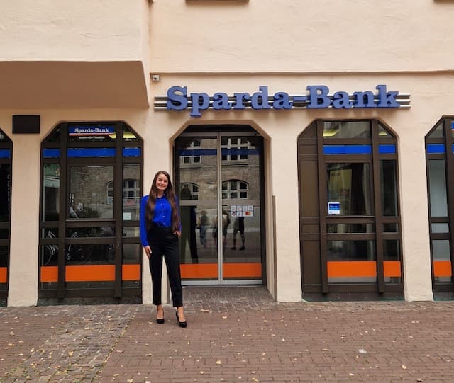 Eine Frau in blauer Bluse steht lächelnd vor einem Sparda-Bank-Gebäude mit großen Fenstern und Ziegelsteinpflaster.