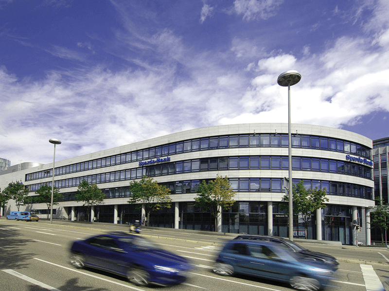 Modernes Bürogebäude mit geschwungener Fassade und großen Fenstern, gelegen an einer belebten Straße mit vorbeifahrenden Autos unter blauem Himmel.