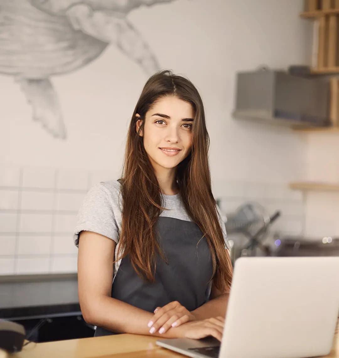 Eine junge Frau steht an einer Theke mit Schürze. Sie hat einen Laptop vor sich stehen.