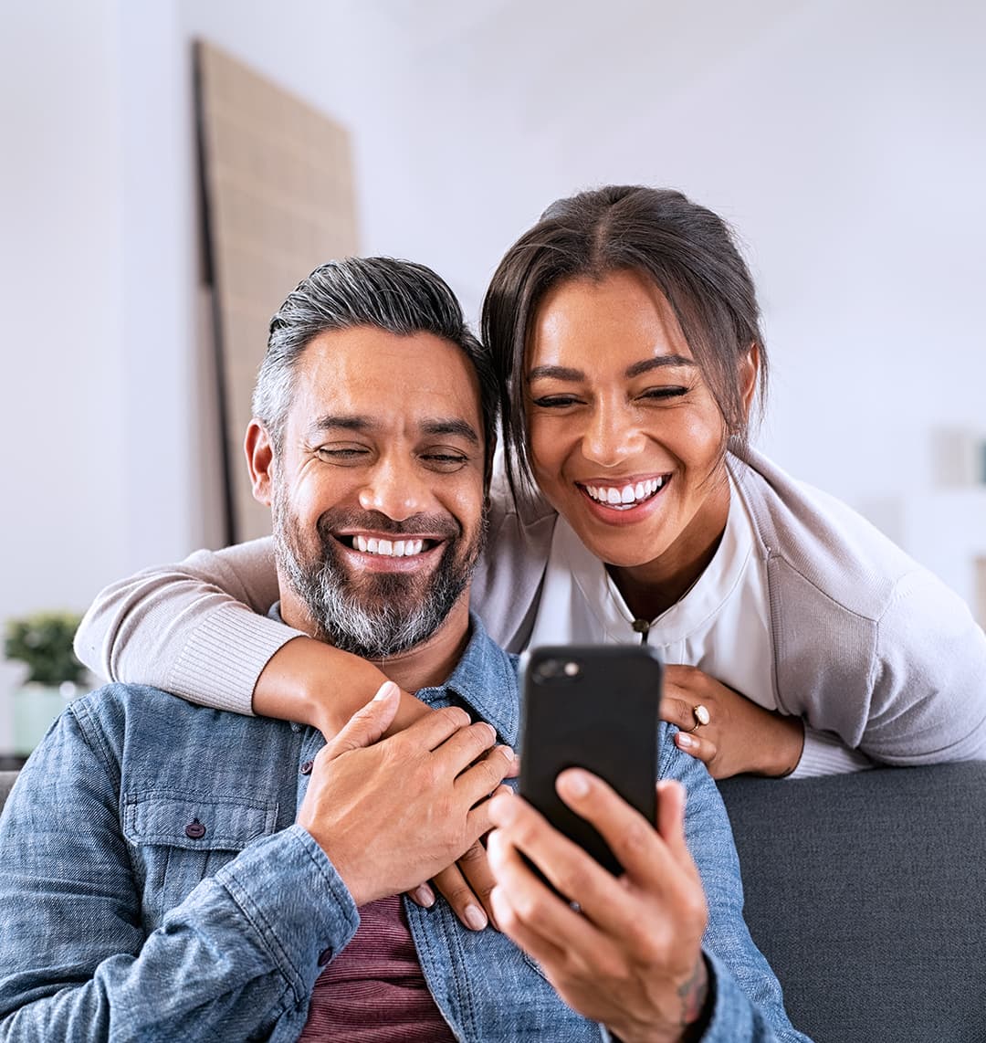 Ein Mann mittleren Alters sitzt auf einem Sofa. Eine Frau steht hinter ihm und umarmt ihn von hinten, er hält ihre Hand. Beide schauen glücklich in das Smartphone in seiner Hand.