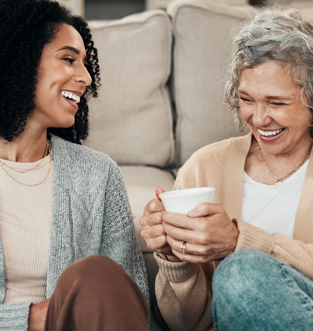 Eine jüngere Frau und eine Frau im mittleren Alter sitzen gemeinsam auf dem Boden vor einem Sofa und trinken Kaffee. Beide lachen herzlich.