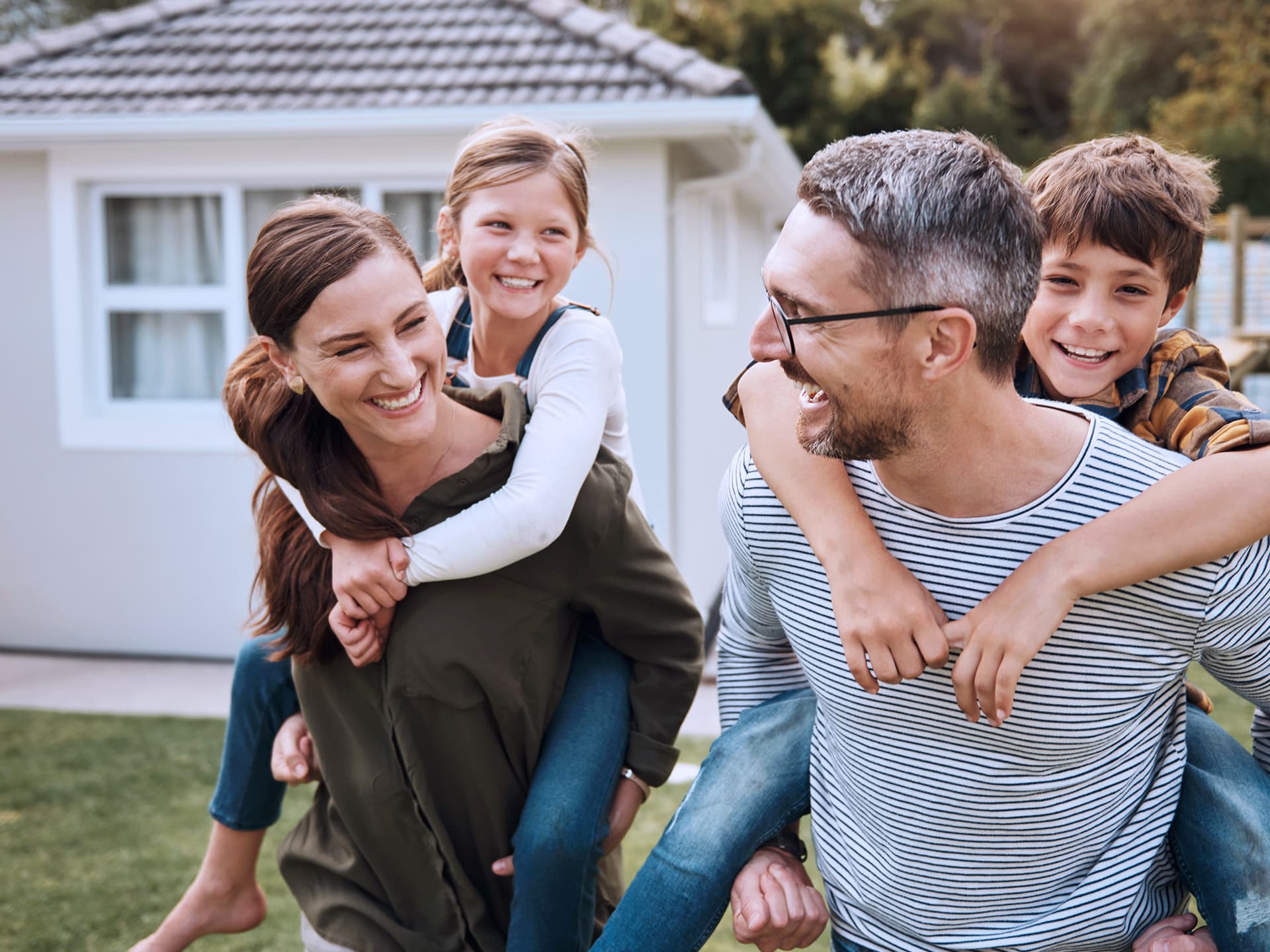 Ein Mann und eine Frau stehen vor einem Haus und tragen jeweils ein Kind huckepack. Alle lachen, der Mann und die Frau schauen sich dabei an.