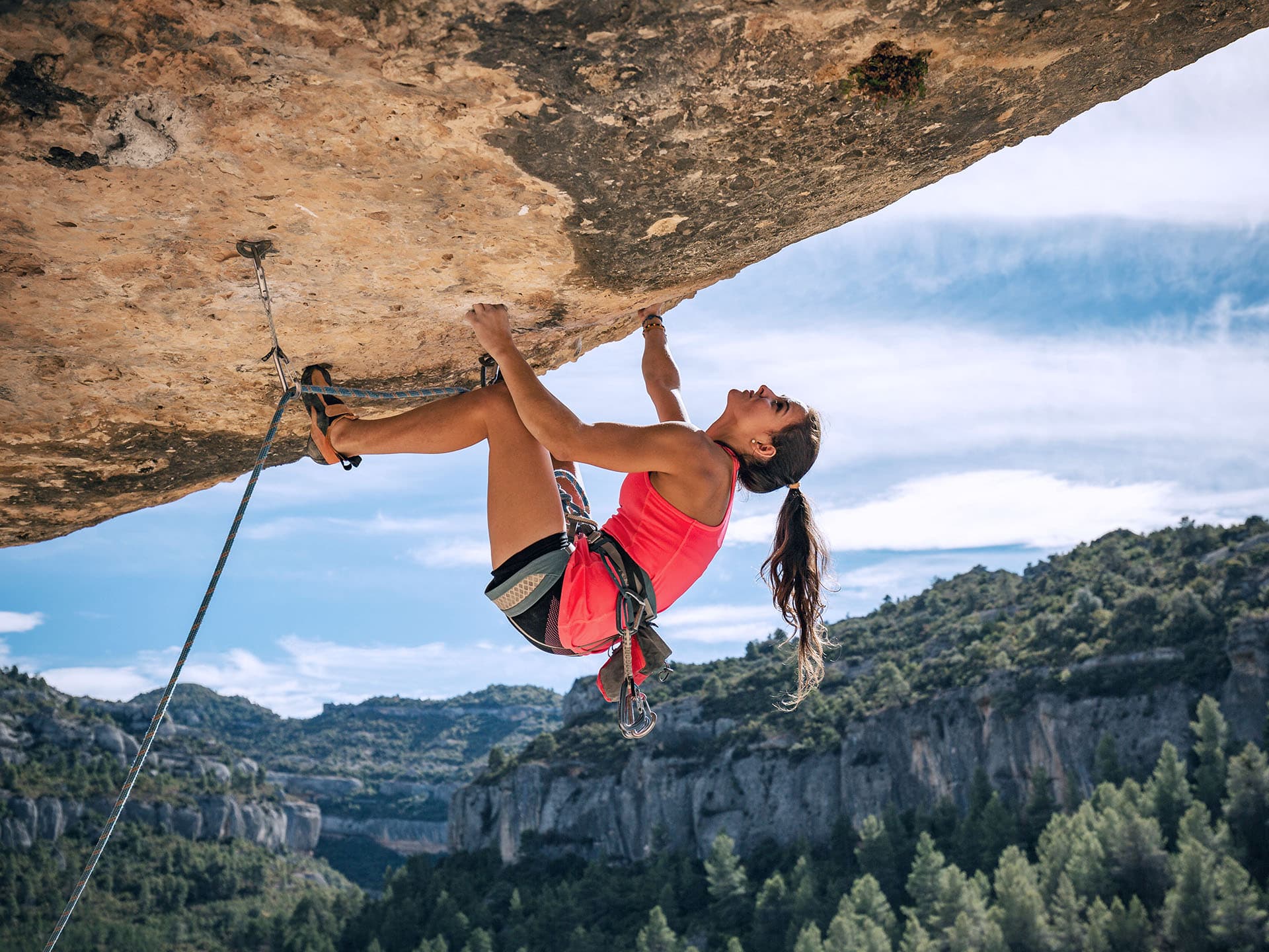 Eine junge Frau klettert an einer Felswand mit Überhang. Im Hintergrund sind weitere Berge zu sehen.